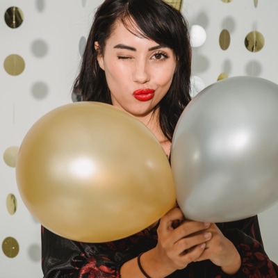 woman wearing bright red lipstick winks at camera while screwing up her mouth; she is wearing a gold shiny party hat and holding gold and silver balloons; there are gold and silver streamers in the background