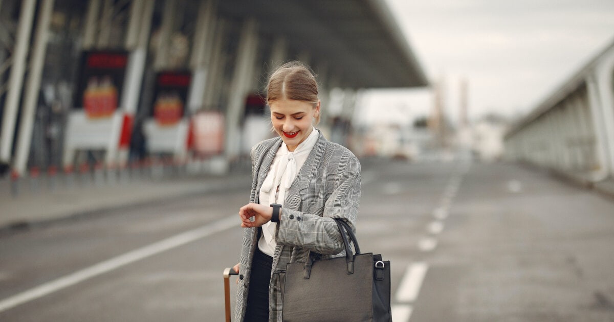 a young professional businesswoman looks at her watch outside an airport while holding her briefcase and a rolling bag; she looks good after a long flight