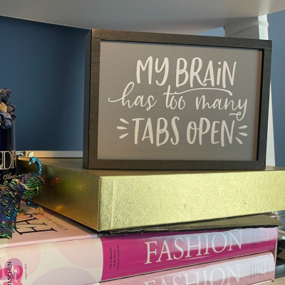 sign reads "My Brain Has Too Many Tabs Open"; the sign sits on top of a gold box on top of two books that read FASHION; in the background is a small metal peacock figurine and a blue mug with paper flowers inside