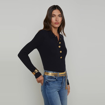 A woman wearing a black polo with gold buttons, blue jeans, a gold bangle, and gold metal belt