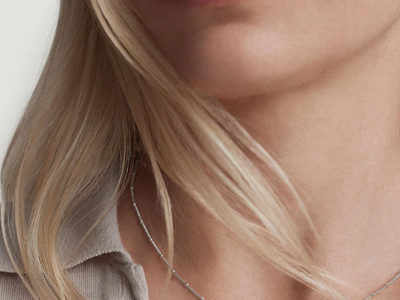blonde woman wears very thin sterling silver necklace with tiny beads as stations; she is also wearing a beige polo sweater