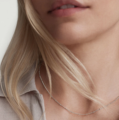 blonde woman wears very thin sterling silver necklace with tiny beads as stations; she is also wearing a beige polo sweater
