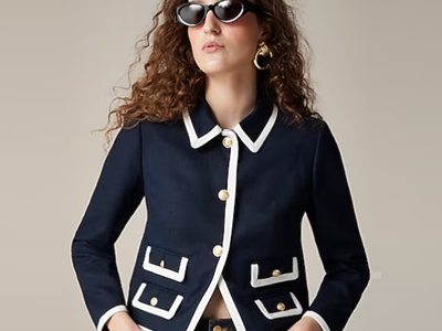 A woman wearing a navy jacket with white trim, sunglasses, and jeans