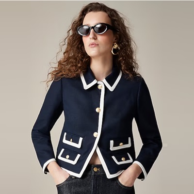 A woman wearing a navy jacket with white trim, sunglasses, and jeans