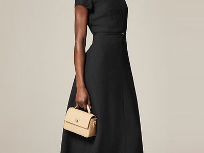 A woman wearing a midi black dress with black heels and a cream-colored handbag