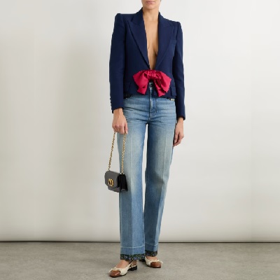 A woman wearing a blue blazer with red ribbon and acid wash denim pants with white sandals and black handbag