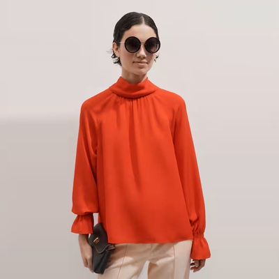 A woman wearing a red-orange blouse, off-white pants, and carrying a black clutch