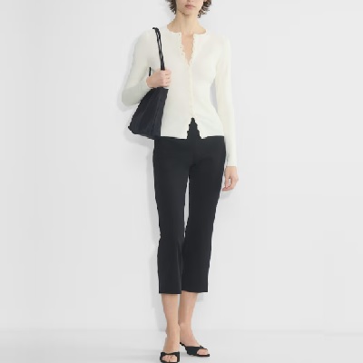 A woman wearing a white long sleeve cardigan top and black pants with black open sandals and black shoulder bag
