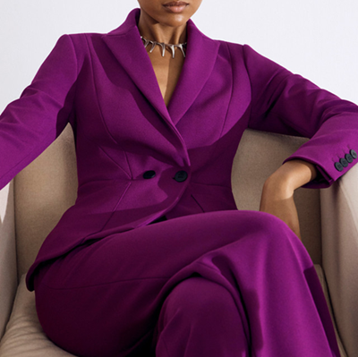 Black model wears vivid berry suit with double breasted blazer; she sits in a chair with her legs crossed
