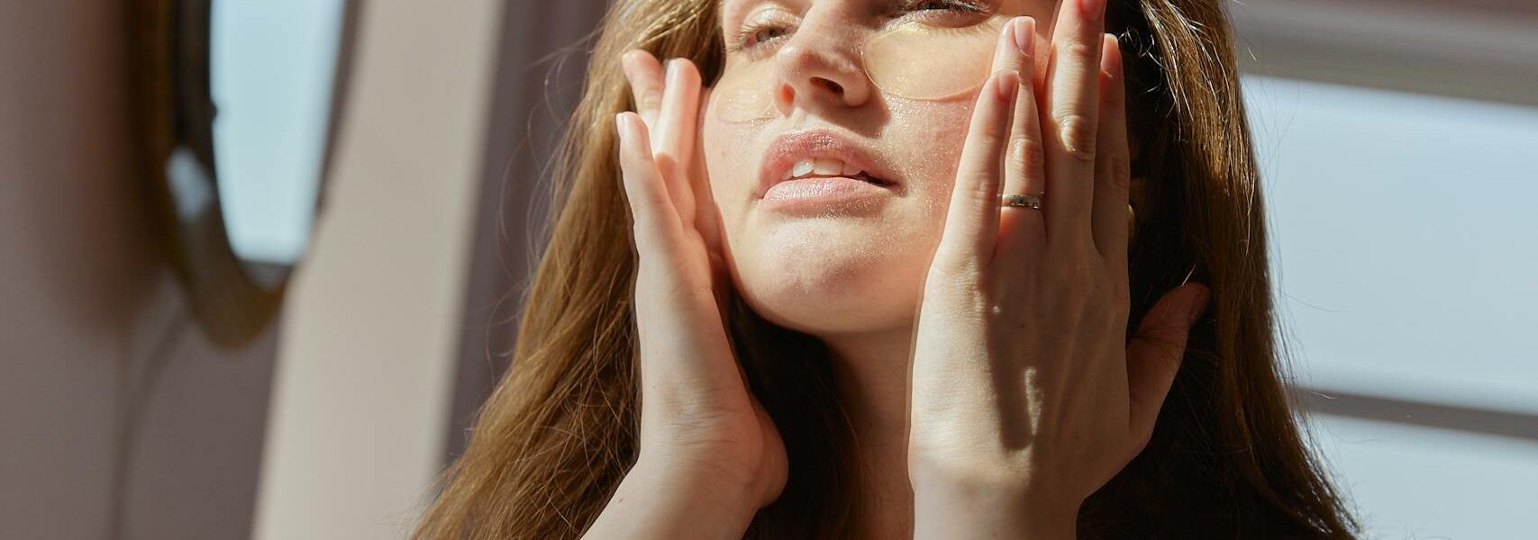 Woman in white tank top applies skincare products while enjoying the sunlight in her home.