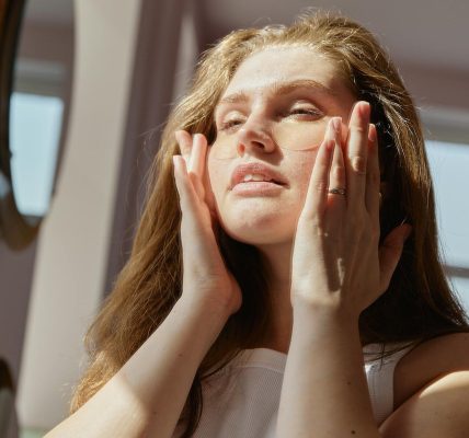 Woman in white tank top applies skincare products while enjoying the sunlight in her home.