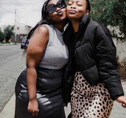 One plussize friend and one straight size friend share a playful moment outdoors, highlighting friendship and joy in an urban setting.