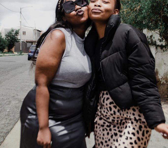 One plussize friend and one straight size friend share a playful moment outdoors, highlighting friendship and joy in an urban setting.