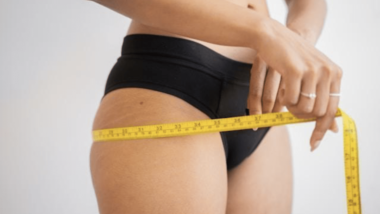 A woman in black panties measuring her hips with yellow measuring tape.