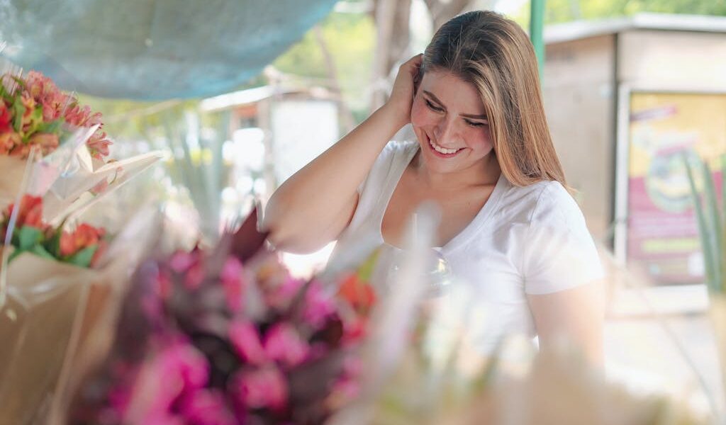 A happy woman in a white t-shirt enjoys browsing vibrant bouquets at a flower market.