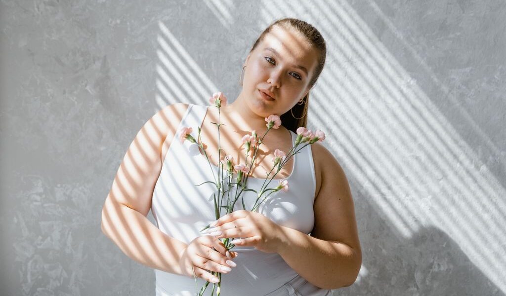 plus size woman in tank top holding flowers with sunlight filtering through blinds. 8433431 scaled